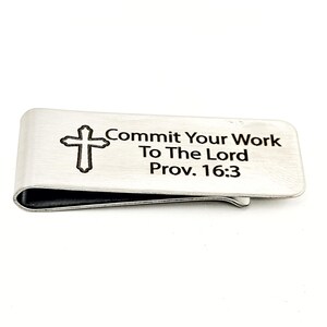 Könnte beinhalten: Ein silberfarbener Geldclip mit einem Kreuz und dem Text "Commit Your Work To The Lord Prov. 16:3". Der Clip hat ein gebogenes Design und eine schwarze Innenseite.