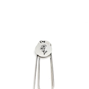 May include: Silver bookmark with a small flower design engraved on a round charm. The bookmark is made of metal and has a simple, elegant design.