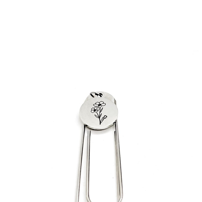 May include: Silver bookmark with a small flower design engraved on a round charm. The bookmark is made of metal and has a simple, elegant design.