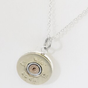 Puede incluir: Un collar de cadena de plata con un colgante redondo hecho de una vaina de escopeta de calibre 20. La vaina est&aacute; marcada con "WIN" y "HS" en el lateral.