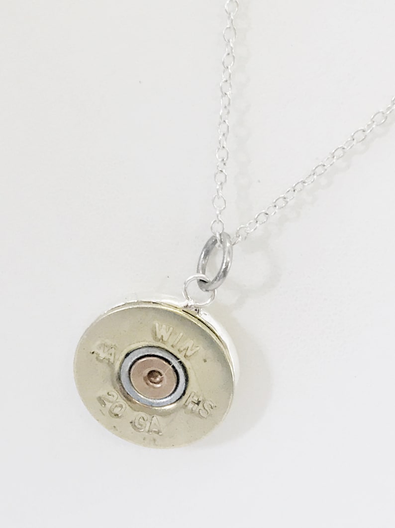 Puede incluir: Un collar de cadena de plata con un colgante redondo hecho de una vaina de escopeta de calibre 20. La vaina est&aacute; marcada con "WIN" y "HS" en el lateral.