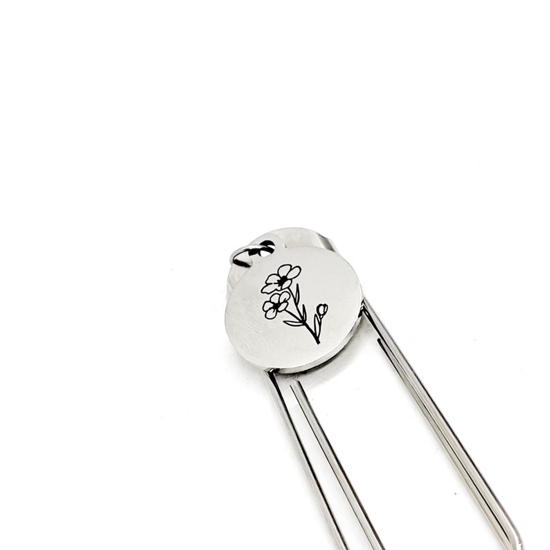 May include: Silver bookmark with a delicate flower design engraved on a round charm. The bookmark has a simple, elegant design.