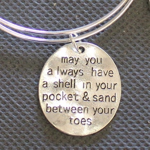 Może przedstawiać: Srebrna bransoletka z okrągłym wisiorkiem, na kt&oacute;rym napisano "may you always have a shell in your pocket & sand between your toes".