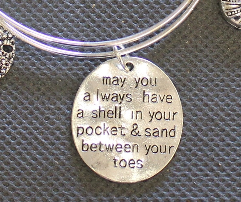 Może przedstawiać: Srebrna bransoletka z okrągłym wisiorkiem, na kt&oacute;rym napisano "may you always have a shell in your pocket & sand between your toes".