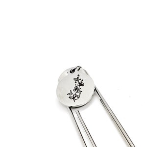 May include: Silver bookmark with a floral design engraved on a round charm. The bookmark has two metal prongs.