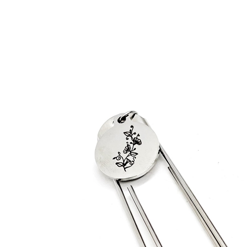 May include: Silver bookmark with a floral design engraved on a round charm. The bookmark has two metal prongs.
