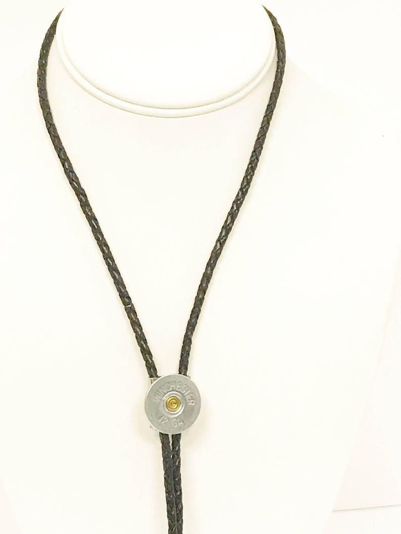 Puede incluir: Un corbat&iacute;n bolo de cuero negro con un colgante de plata de una vaina de cartucho de escopeta. La vaina tiene el texto "WINCHESTER 12 GA" en ella.