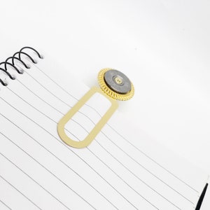 May include: A gold bookmark with a round, silver, and gold decorative top. The bookmark is designed to hold a page in a book.