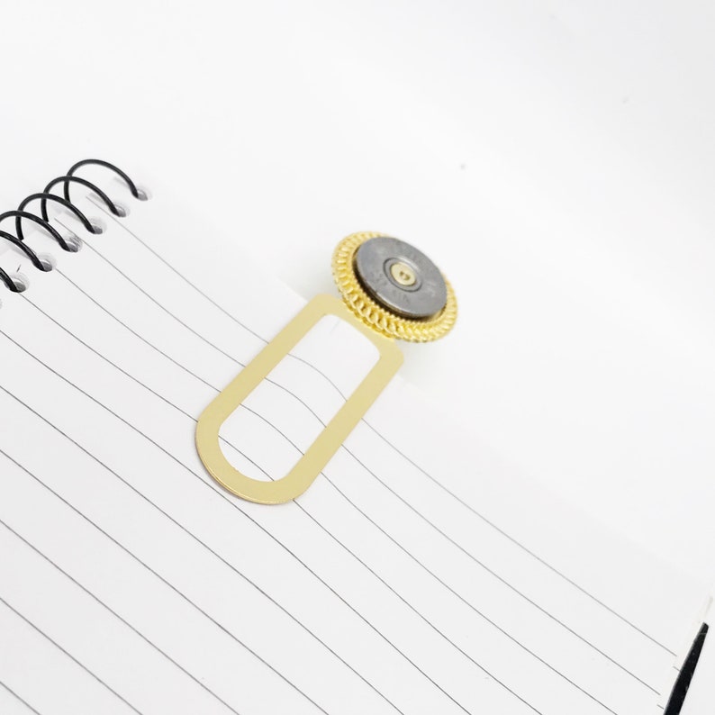 May include: A gold bookmark with a round, silver, and gold decorative top. The bookmark is designed to hold a page in a book.