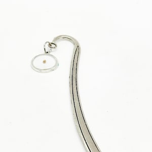 May include: A silver bookmark with a curved design and a small, circular charm. The charm contains a mustard seed. The bookmark has a textured pattern along its curved edge. The background is plain white.