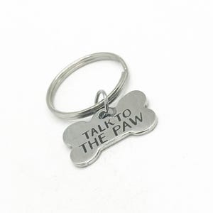 Op de afbeelding: Zilveren hondenbot sleutelhanger met de tekst "Talk to the Paw" erop gegraveerd.