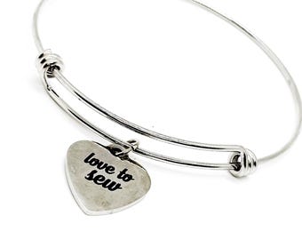Op de afbeelding: Een zilveren, verstelbare armband met een hartvormige bedel waarop "love to sew" staat.
