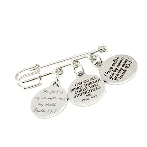 May include: Silver safety pin with four round charms. Each charm has a different bible verse engraved on it. The verses are: "The Lord is my strength and my shield. Psalm 28:7", "I can do all things through Christ who strengthens me. Phil 4:13", and "I have called you by name; you are mine. Isaiah 43:1".