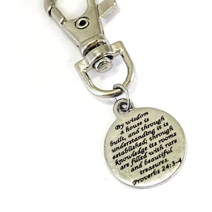 May include: Silver-coloured keychain with a round charm. The charm has the text "By wisdom a house is built... Proverbs 24:3-4" in black lettering. The keychain has a clasp for easy attachment.