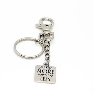 May include: Silver-toned keychain with a lobster clasp, key ring, and a small square charm. The charm is engraved with the words "pray MORE worry LESS" in a simple, elegant font. The keychain is designed for everyday use.