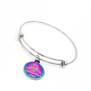 May include: A silver-toned adjustable bangle bracelet with a circular charm. The charm has a rainbow iridescent finish and features the text "be a rainbow in someone else's cloud" in a script font.