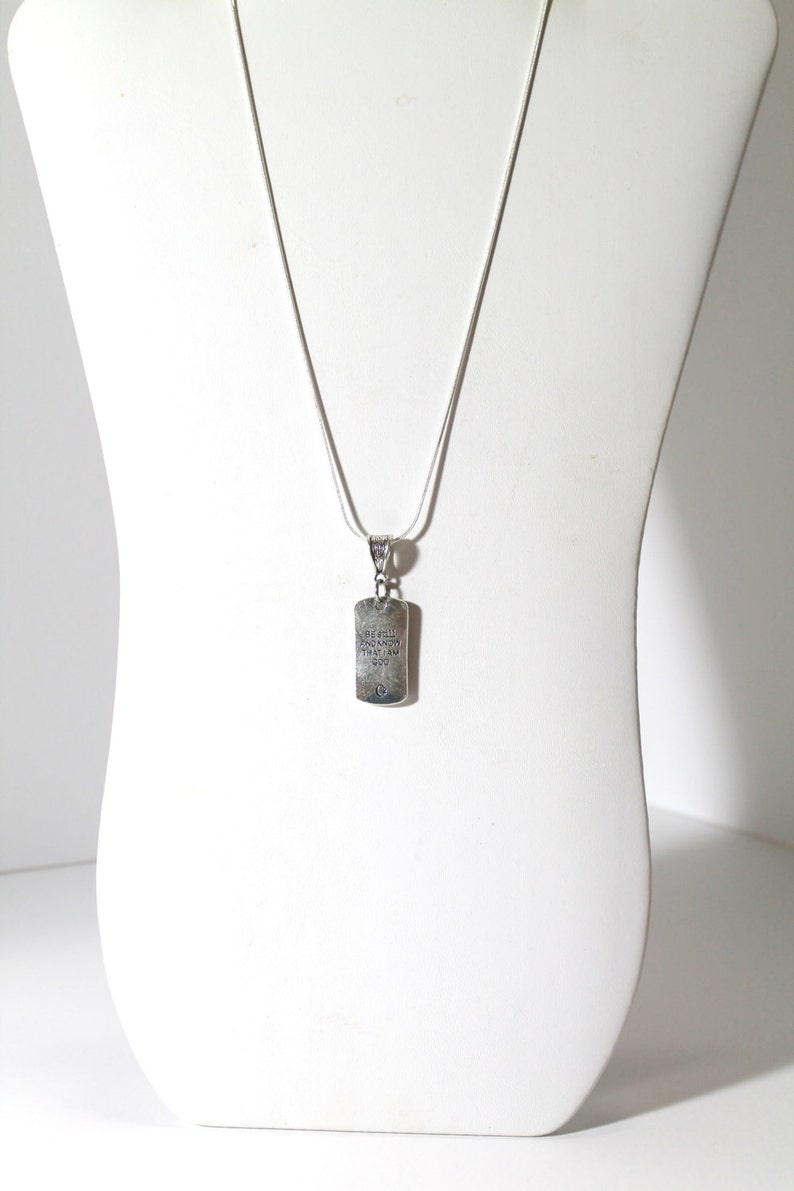 May include: A silver chain necklace with a silver pendant. The pendant is rectangular and has the words "Be still and know that I am God" engraved on it.