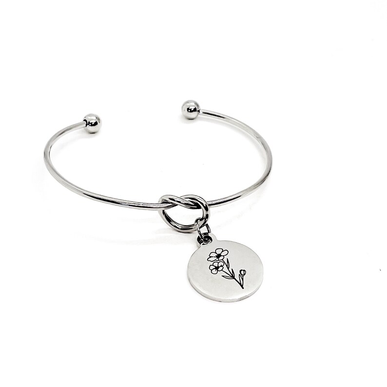May include: Silver bangle bracelet with a knotted design and a charm featuring a delicate flower illustration.