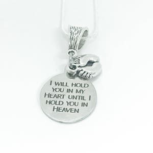 May include: A silver-colored pendant necklace with a round charm. The charm has the engraved text: "I WILL HOLD YOU IN MY HEART UNTIL I HOLD YOU IN HEAVEN." The pendant features a detailed design and is suspended from a delicate silver chain.