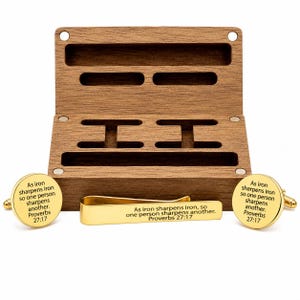 May include: A set of two round gold-tone metal cufflinks and a matching gold-tone tie bar, each engraved with the text 'As iron sharpens iron, so one person sharpens another. Proverbs 27:17', presented in a rectangular brown wooden storage box.