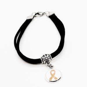 May include: Black suede bracelet with a silver charm featuring a gold ribbon. The charm is attached to the bracelet with a silver bead.