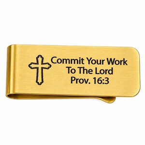May include: A gold-coloured money clip featuring a cross and the inscription "Commit Your Work To The Lord Prov. 16:3". The clip has a curved design and is made of shiny metal.