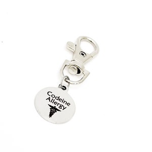 May include: Silver keychain with a round tag that says "Codeine Allergy" with a medical symbol. The keychain has a silver clip.