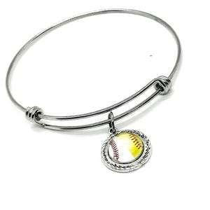 May include: A silver-toned bangle bracelet with a charm featuring a softball design. The charm is round, with a yellow and white softball image and a braided silver border. The bracelet is adjustable and has a simple, elegant design.