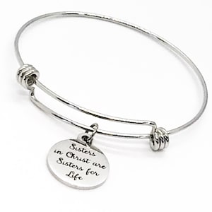 Puede incluir: Una pulsera de brazalete plateada con un dije redondo. El dije está grabado con las palabras "Sisters in Christ are Sisters for Life". La pulsera es de un material metálico brillante.