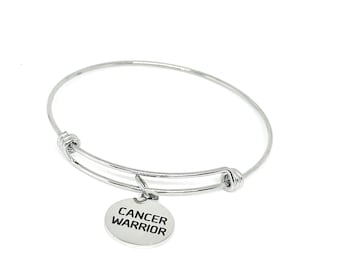 May include: Silver-toned bangle bracelet with a round charm. The charm is engraved with the words "CANCER WARRIOR" in black, centered on the charm. The bracelet is made of a shiny, metallic material and is set against a white background.