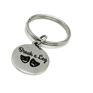 May include: A silver-colored keychain with a round charm. The charm has the phrase "Break a Leg" in black script, along with two theater masks. The keychain ring is attached to the charm with a small loop.