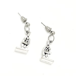 May include: Silver-colored microscope earrings. Each earring features a miniature microscope charm dangling from a post. The microscope charms have detailed designs, including the base, stage, and ocular lens. The earrings are designed for pierced ears.