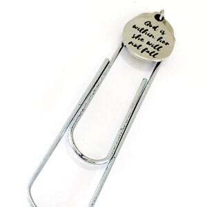 May include: A silver paperclip bookmark with a round charm attached. The charm has the words "God is within her she will not fall" engraved on it.