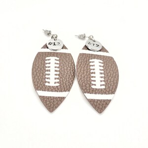 May include: A pair of brown leather football-shaped earrings with white stitching and a silver charm with the number "#12" on each earring.