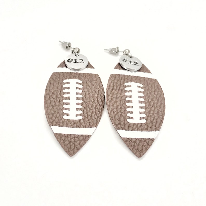 May include: A pair of brown leather football-shaped earrings with white stitching and a silver charm with the number "#12" on each earring.