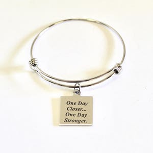 May include: Silver bangle bracelet with a charm that reads "One Day Closer... One Day Stronger."