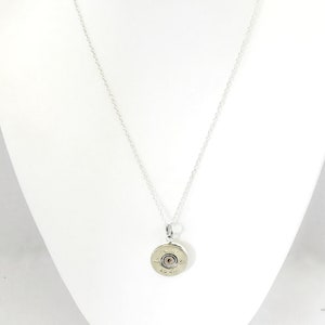 Puede incluir: Un collar de cadena de plata con un colgante hecho de una vaina de cartucho de escopeta. La vaina es de color dorado y tiene un peque&ntilde;o centro redondo de plata.