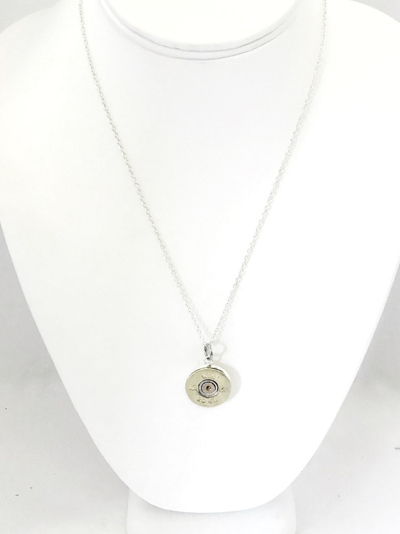 Puede incluir: Un collar de cadena de plata con un colgante hecho de una vaina de cartucho de escopeta. La vaina es de color dorado y tiene un peque&ntilde;o centro redondo de plata.