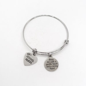 May include: Silver expandable bracelet with two charms. One charm is a heart with the text "School Nurse" and a small image of a bandage. The other charm is a circle with the text "Bless me with a gentle touch and a caring heart".