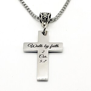 May include: A silver-colored cross pendant with the inscription "Walk by faith 2 Cor. 5:7" is suspended from a matching chain. The cross has a simple, rectangular design with rounded edges. The chain is a linked style.