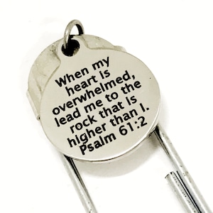 May include: A silver-colored bookmark with a circular charm. The charm has the text "When my heart is overwhelmed, lead me to the rock that is higher than I. Psalm 61:2" in black lettering. The bookmark is on a white background.