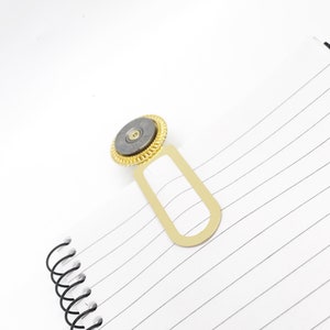May include: Gold bookmark with a round, gray metal design in the center. The bookmark has a rectangular shape with a rounded top and bottom.
