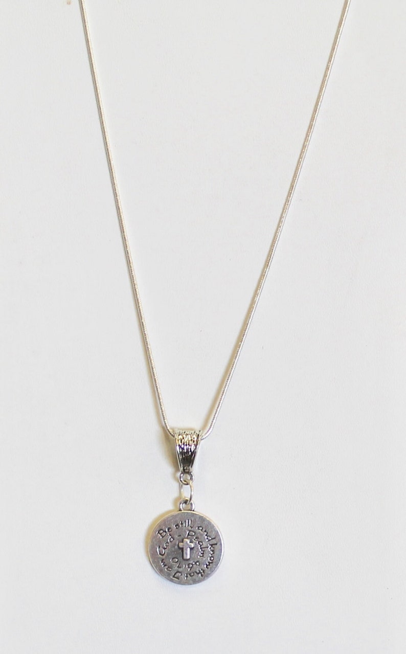 May include: A silver chain necklace with a round pendant that reads "Be still and know that I am God". The pendant features a small cross in the center.