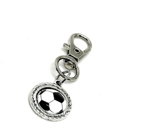 May include: A silver-colored soccer ball charm with a black and white design, attached to a metal clip. The charm is round with a textured border. The clip is a standard clasp.