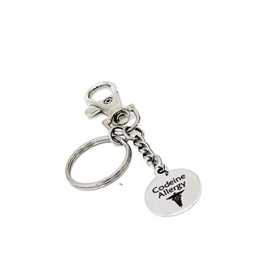 May include: Silver keyring with a round tag that says "Codeine Allergy" with a medical symbol. The keyring has a split ring and a lobster clasp.