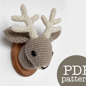 Könnte beinhalten: Gehäkelte Hirschkopf-Wanddekoration aus braunem und weißem Garn. Der Hirschkopf ist auf einer Holzplatte montiert. Der Text "PDF pattern" befindet sich auf einem braunen Kreis.