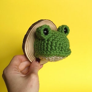May include: A green crocheted frog head mounted on a wooden slice. The frog has black eyes and a beige crocheted underside.