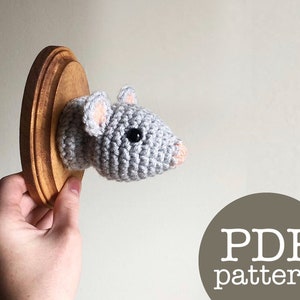 May include: A crocheted gray mouse head mounted on a wooden plaque. The mouse has a pink nose and black eyes. The plaque is brown and has a natural wood finish. The text "PDF pattern" is written in white on a brown circle.