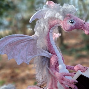 May include: A pink plush dragon with white fur and wings is reading a book. The dragon is wearing glasses and has a friendly expression.