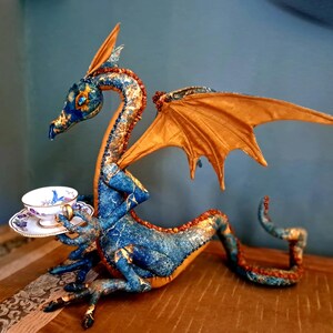 Puede incluir: Una figura decorativa de dragón con un acabado texturizado en azul y dorado, grandes alas de color ámbar y una cola larga. El dragón sostiene en sus garras una pequeña taza de té y un plato de porcelana blanca con motivos florales azules.